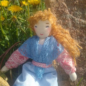 May include: A handmade fabric doll with curly ginger hair, wearing a light blue dress with a lace shawl and pink patterned sleeves. The doll has a simple embroidered face with rosy cheeks and is positioned against a natural outdoor background.