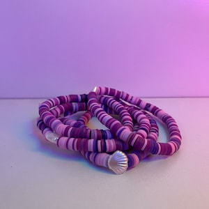 May include: A collection of purple and lavender beaded bracelets. The bracelets are made of small, flat, round beads in varying shades of purple and lavender. Each bracelet has a silver seashell charm. The bracelets are stacked together.