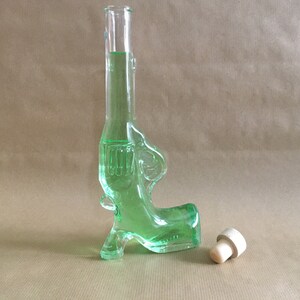 Könnte beinhalten: Eine grüne Glasflasche in Form einer Pistole mit einem Korkstopfen. Die Flasche ist mit einer grünen Flüssigkeit gefüllt.