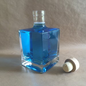 Può includere: Una bottiglia di vetro trasparente, di forma quadrata, riempita di liquido blu brillante, accanto a un tappo di sughero. La bottiglia ha un design angolare unico. Il tappo è in legno chiaro. Lo sfondo è marrone neutro.
