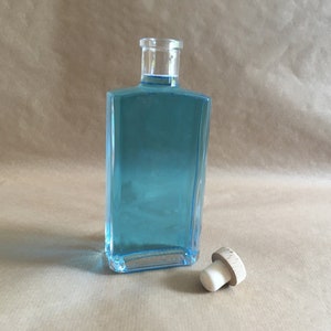 May include: A clear glass bottle with a blue liquid inside and a wooden cork stopper. The bottle is rectangular and has a square base.