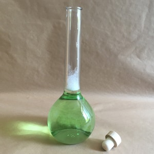 Puede incluir: Una botella de vidrio transparente con fondo redondo y cuello largo. La botella está llena de un líquido verde y tiene una espuma blanca en la parte superior. Un tapón de corcho de madera está al lado de la botella.