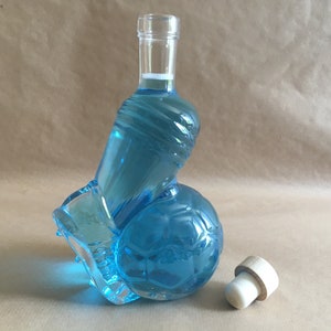 Op de afbeelding: Een blauwe glazen fles in de vorm van een voetbal met een kurken stop. De fles is gevuld met een lichtblauwe vloeistof.