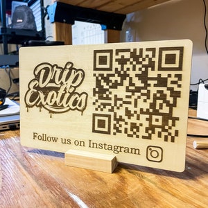 May include: A light-colored wooden sign with the words "Drip Exotics" in a stylized font, a QR code, and the text "Follow us on Instagram" with the Instagram logo. The sign is propped up by a small wooden stand.
