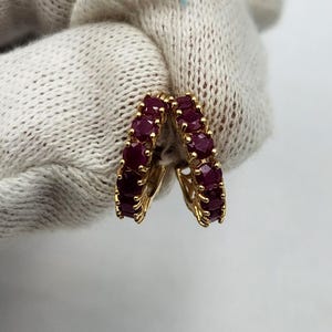 May include: A pair of gold hoop earrings featuring a row of round, deep red rubies set along the front curve of each piece. The earrings are designed with a hinged closure mechanism, showcasing a polished gold metal finish and vibrant gemstones.