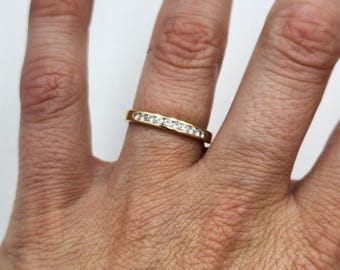 May include: A gold band ring with a row of small, clear gemstones set across the top. The ring is on a finger, and the background is a neutral color.