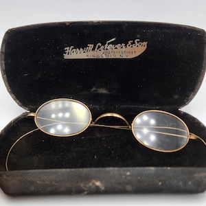 May include: Vintage oval eyeglasses with gold-tone frames rest inside an open black velvet case. The case is embossed with the text "Harry R. Lefever & Son, 292 Fair Street, Kingston NY."