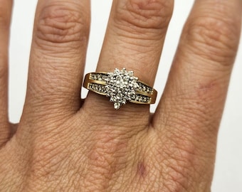 May include: A gold ring featuring a central cluster of small round clear stones arranged in a floral shape, with additional small clear stones set along the split shank of the band. The metal has a polished yellow gold finish throughout.