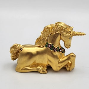 May include: A gold unicorn figurine with a jeweled flower garland around its neck. The unicorn is lying down with its head turned to the side and its horn pointing forward.