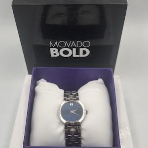 May include: A Movado Bold watch with a blue dial and silver-tone stainless steel band, presented in a black box with the brand name. The watch is displayed on a white pillow inside a white and purple box.