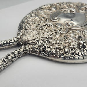 May include: Ornate silver hand mirror with a floral design. The mirror features a round shape with a handle, and is covered in detailed carvings of flowers and leaves. The mirror's surface is reflective.