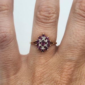 May include: A gold ring featuring a cluster of gemstones. The ring's centerpiece is a marquise-cut, deep red gemstone, surrounded by smaller, round, red gemstones and interspersed with white gemstones. The ring is set on a gold band.