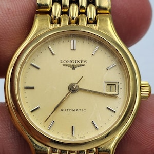 May include: A gold-tone Longines automatic wristwatch featuring a round champagne-colored dial with gold-tone hour markers, a date window at the three o'clock position, and a multi-link metal bracelet with a polished finish and a secure clasp.