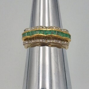 May include: A gold band ring with a row of emerald green square-shaped stones set in a gold band, surrounded by a row of smaller round white stones.