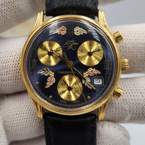 May include: Gold-toned chronograph watch with a navy blue dial and black leather strap. The watch features gold, rose gold, and silver-toned leaf accents. The dial includes subdials, Roman numerals, and the date.