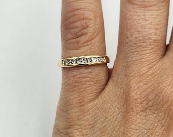 May include: A gold ring with a row of small, square-cut diamonds. The ring is set in a yellow gold band and is worn on a finger.