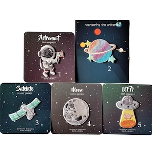 May include: Five embroidered iron-on patches with a space theme. The patches are on a black background with white text that says "From My Universe". The patches are numbered 1 through 5. The patches depict an astronaut, a planet, a satellite, a moon, and a UFO.