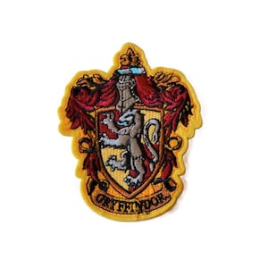 May include: An embroidered patch featuring a shield with a gold background and a gray lion rampant. The shield is surrounded by red and gold decorative flourishes, topped with a helmet, and includes the text GRYFFINDOR at the bottom.