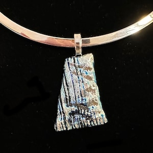 Puede incluir: Un collar plateado con un colgante. El colgante tiene forma rectangular con una superficie texturizada e iridiscente en tonos azules, plateados y verdes. El collar se muestra sobre un fondo negro.