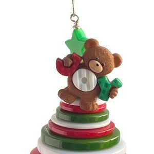 Puede incluir: Un adorno de árbol de Navidad con forma de pila de botones coloridos con un oso de peluche marrón que sostiene las letras "JOY" en la parte superior.