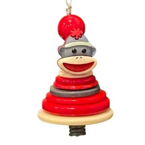 May include: A red and gray sock monkey ornament with a red ball on top. The monkey is smiling and has a stack of red and gray rings around its neck.