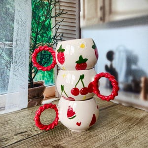 Puede incluir: Una pila de tres tazas de cerámica blanca con asas rojas. Cada taza presenta un diseño de fruta diferente: fresas, cerezas y sandía.