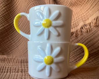 May include: Two white ceramic mugs with yellow handles and a raised daisy design. The daisies have white petals and yellow centers.