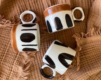 Può includere: Tre tazze in ceramica con disegni astratti in bianco e nero. Le tazze hanno bordi e manici marroni.