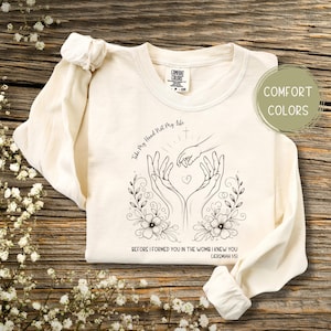 Puede incluir: Una camiseta de manga larga de color crema con un diseño gráfico en blanco y negro de dos manos formando un corazón con una cruz arriba. El texto "Take My Hand Not My Life" está por encima de las manos y el texto "BEFORE I FORMED YOU IN THE WOMB I KNEW YOU (JEREMIAH 1:5)" está debajo de las manos.