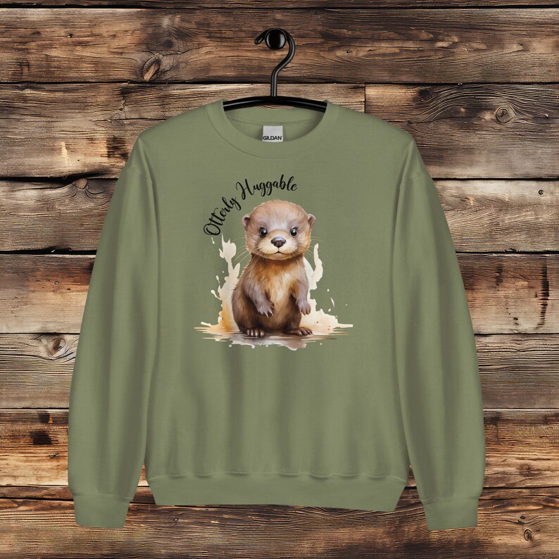 May include: A green sweatshirt with a cute cartoon otter on the front. The otter is surrounded by a watercolor-style splash of color. The text "Otterly Huggable" is written above the otter.