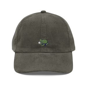 May include: A green corduroy baseball cap with an embroidered green frog holding a white flower.