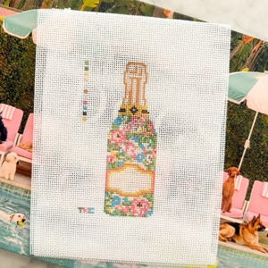 May include: A white needlepoint canvas featuring a central design of a champagne bottle decorated with a colorful floral pattern in shades of pink, blue, and green, including a blank rectangular label area and a small color key on the left side.