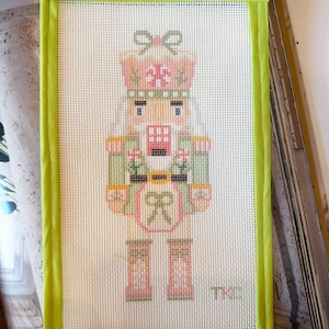 May include: A rectangular needlepoint canvas featuring a nutcracker design in shades of pink, sage green, and white. The nutcracker wears a hat with a bow and has a bow on its chest. The canvas is finished with a bright lime green fabric border.