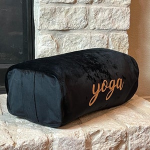 Puede incluir: Un cojín de yoga de terciopelo negro con la palabra "yoga" bordada en oro.