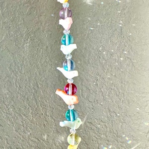 Könnte beinhalten: Ein Kristall-Sonnenfänger mit einem Regenbogen aus Farben und kleinen Vogelanhängern. Der Sonnenfänger hängt an einer Decke und wirft Regenbögen an die Wand.