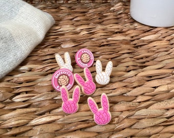 May include: Five pink and white bunny-shaped charms with a brown and white pastry design. The charms have the text "Cafecito & Chisime" on them.
