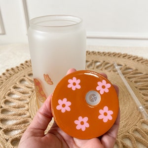 May include: Orange lid with a hole in the center and pink flower designs. The lid is for a glass bottle.