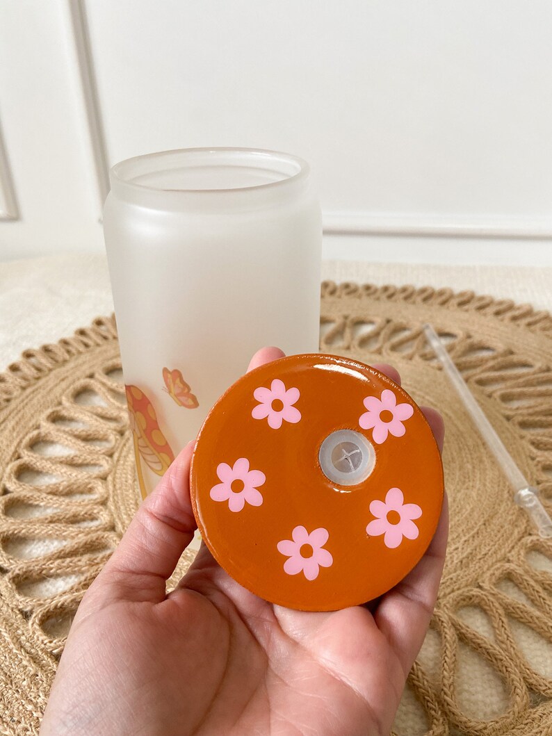 May include: Orange lid with a hole in the center and pink flower designs. The lid is for a glass bottle.