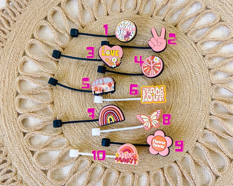 May include: A collection of ten colorful, decorative charms with black elastic bands. The charms feature various designs, including a peace sign, a rainbow, a butterfly, and the words "Love" and "Peace Love". The color palette includes pink, orange, and white.