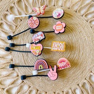 May include: A collection of colorful shoe charms with a retro theme. The charms feature designs like a butterfly, flowers, a peace sign, a rainbow, and the words "Stay Groovy" and "Peace Love". The charms are on black cords.