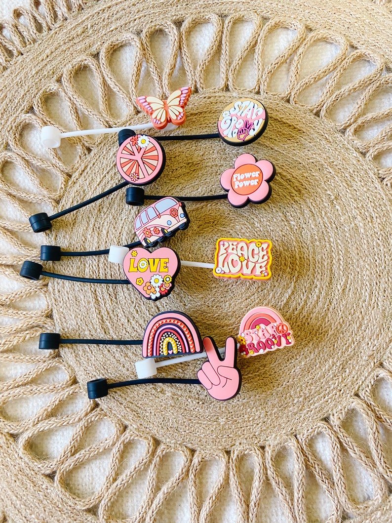 May include: A collection of colorful shoe charms with a retro theme. The charms feature designs like a butterfly, flowers, a peace sign, a rainbow, and the words "Stay Groovy" and "Peace Love". The charms are on black cords.