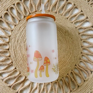 May include: A frosted glass water bottle with a brown lid and a straw. The bottle has a design of yellow mushrooms with red tops and pink flowers.