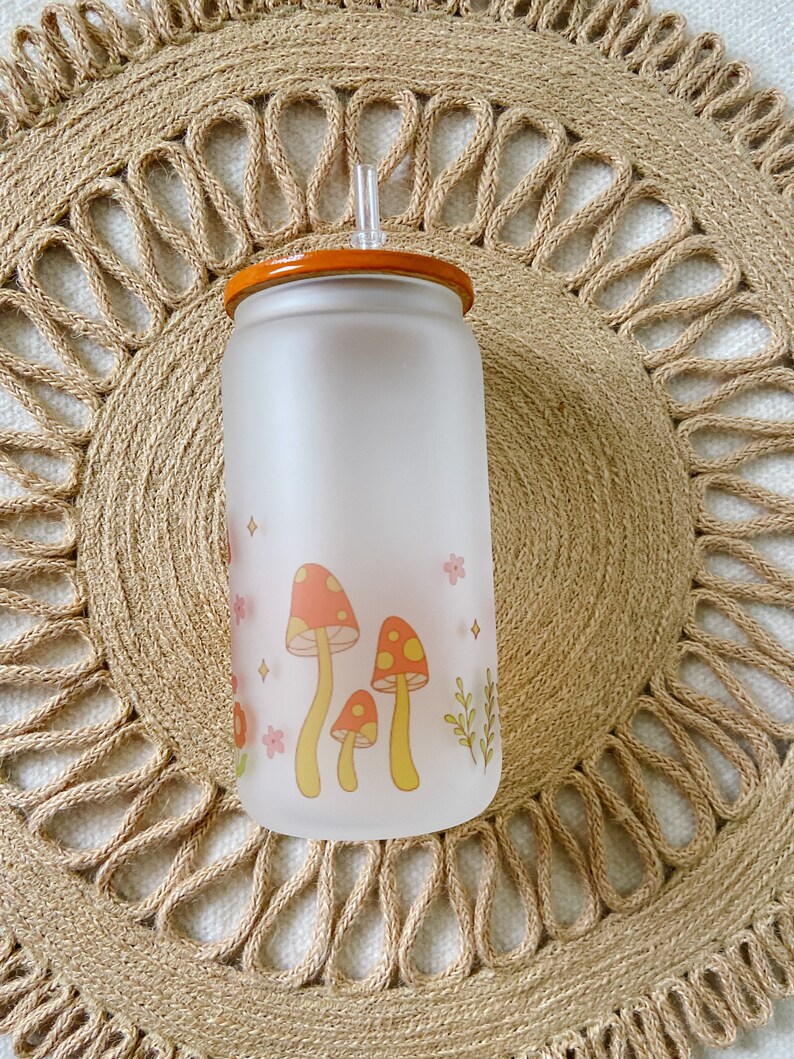 May include: A frosted glass water bottle with a brown lid and a straw. The bottle has a design of yellow mushrooms with red tops and pink flowers.