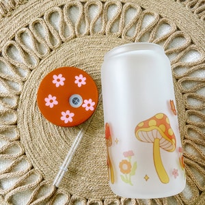 May include: A frosted glass water bottle with a mushroom and flower design. The bottle has a brown lid with pink flowers.
