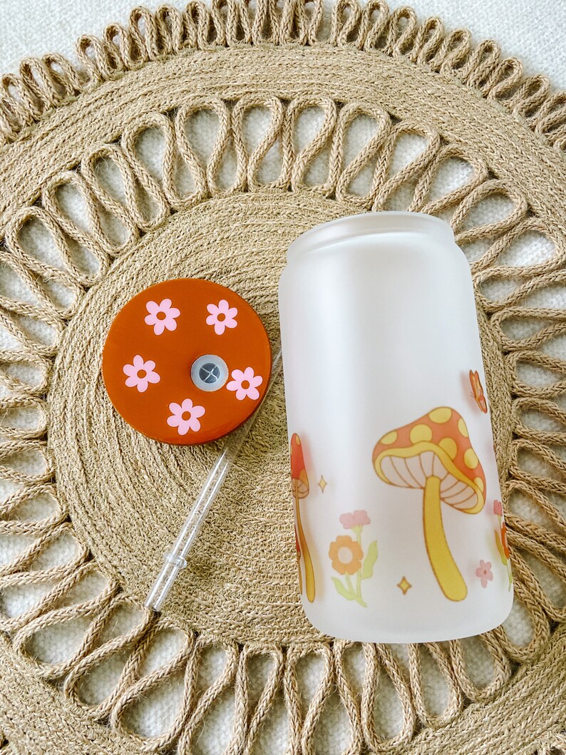 May include: A frosted glass water bottle with a mushroom and flower design. The bottle has a brown lid with pink flowers.