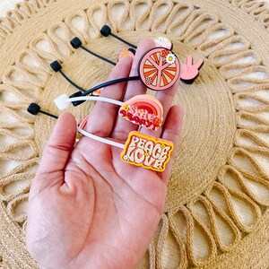 May include: A collection of colorful, retro-themed shoe charms. Designs include a peace sign, a rainbow, and the words "Peace Love" in a groovy font. The charms are attached to black and white rubber bands.