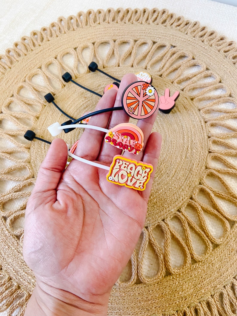 May include: A collection of colorful, retro-themed shoe charms. Designs include a peace sign, a rainbow, and the words "Peace Love" in a groovy font. The charms are attached to black and white rubber bands.