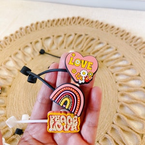 May include: A collection of colorful shoe charms featuring a heart with the word "LOVE", a rainbow, and a sign that says "PEACE LOVE". The charms are in shades of pink, yellow, orange, and red. The charms are on a neutral background.