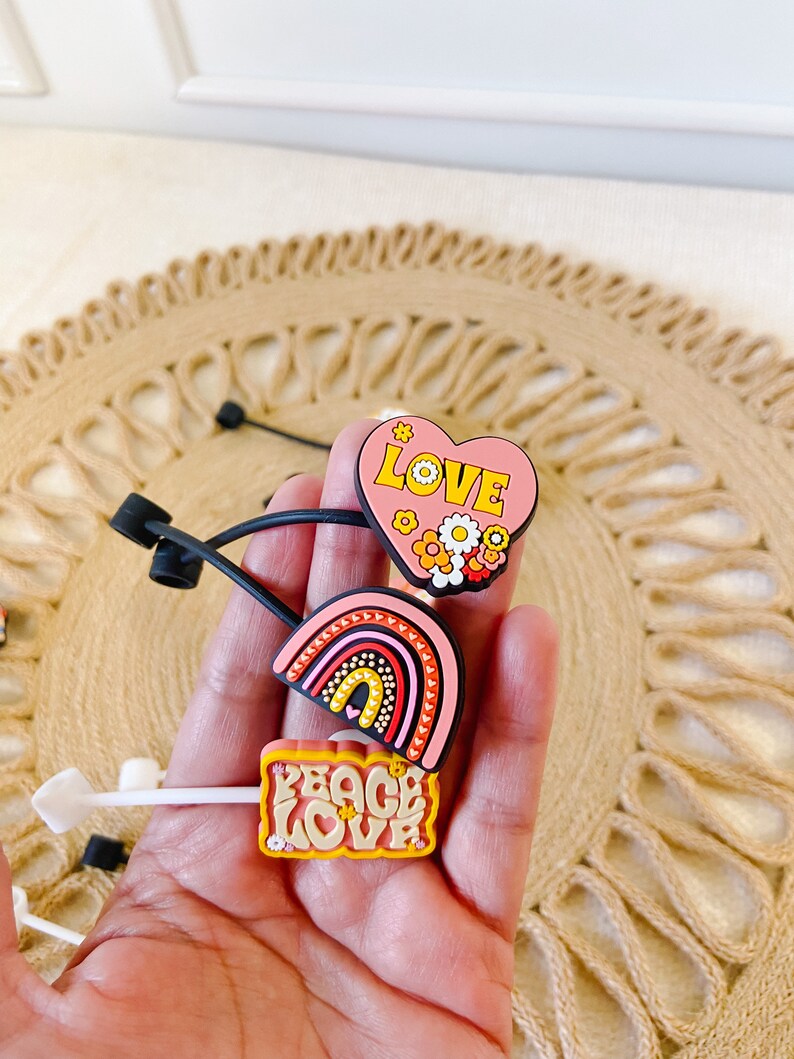 May include: A collection of colorful shoe charms featuring a heart with the word "LOVE", a rainbow, and a sign that says "PEACE LOVE". The charms are in shades of pink, yellow, orange, and red. The charms are on a neutral background.