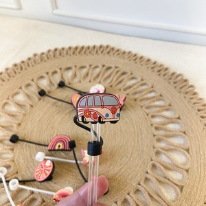 May include: A clear straw with a black silicone ring and a decorative topper featuring a pink and peach-colored retro van with flower accents. Other straw toppers with rainbows, butterflies, and fruit are also visible.
